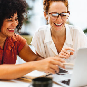 Zwei Frauen sitzen vor einem Laptop, zeigen auf den Bildschirm und sind gut gelaunt.