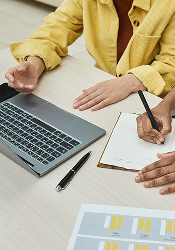 Nahaufnahme auf die Hände zweier Personen, die auf einen Laptop zeigen und Notizen auf einem Schreibblock machen.
