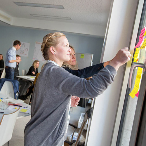 Eine junge Frau klebt während eines Seminars mit einer weiteren Teilnehmerin Post-its an ein Fenster.