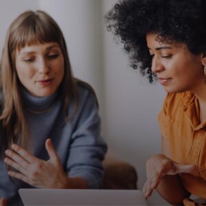 Zwei Frauen sitzen zusammen und diskutieren vor einem Laptop, eine spricht gestikulierend, die andere hört aufmerksam zu.