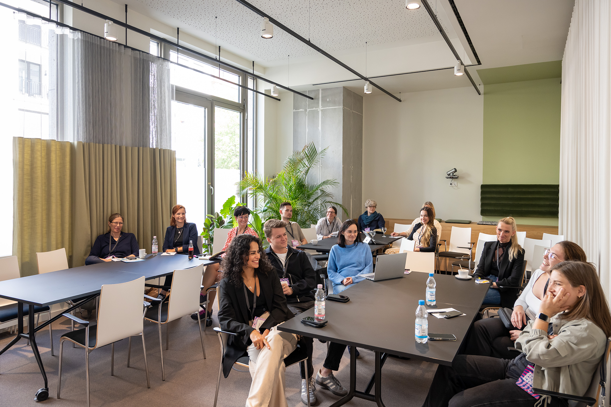 Gruppensitzung in modernem Raum mit Teilnehmern an Tischen, Pflanzen im Hintergrund und großen Fenstern.