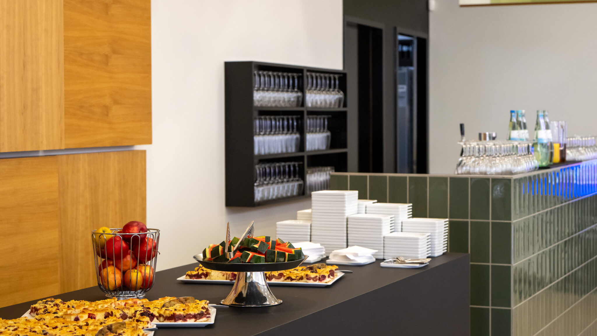 Buffet mit Obst, Snacks und Geschirr in einem modernen Studienzentrum. Im Hintergrund Gläser und eine Bar.