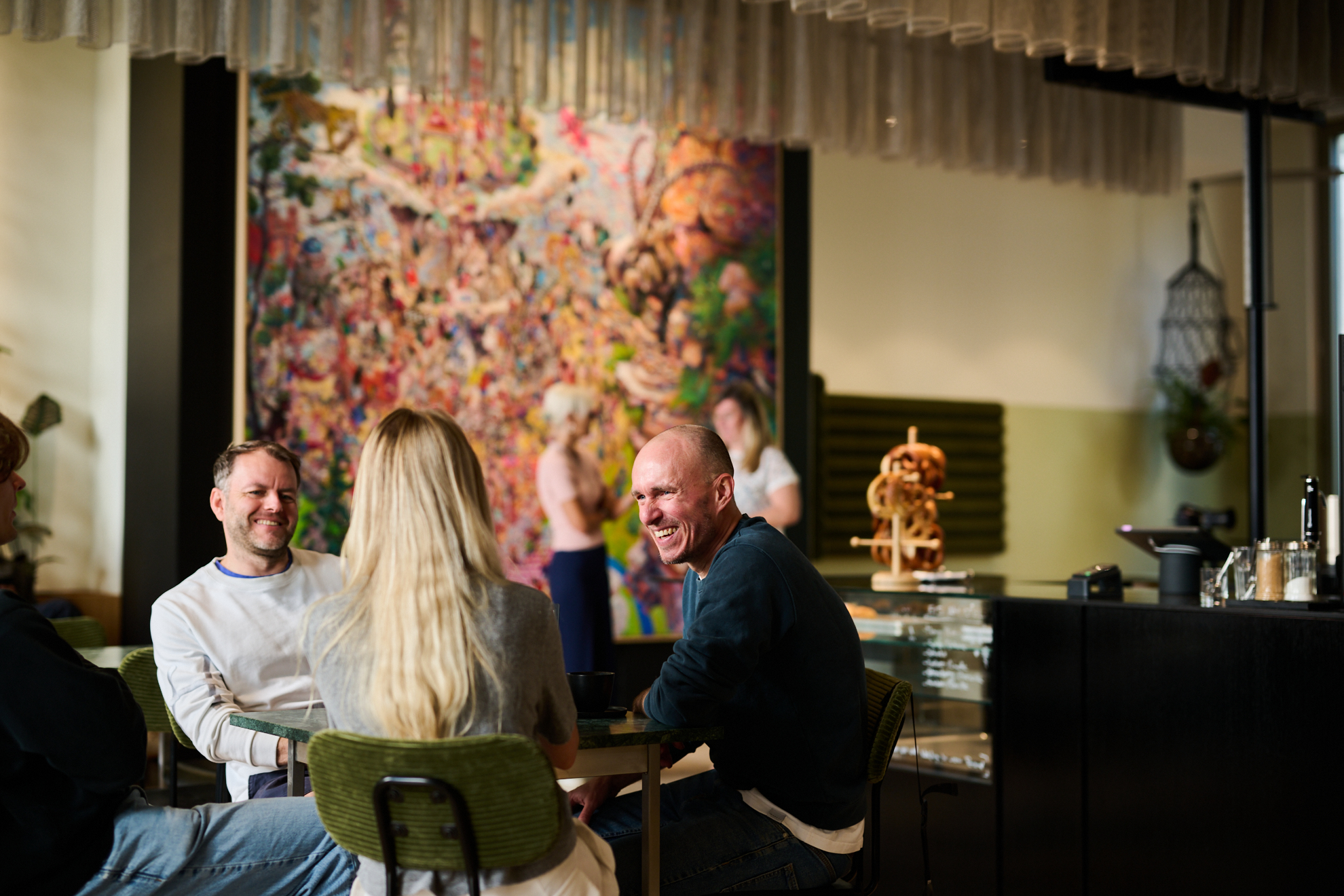 Drei Personen sitzen an einem Tisch in einem modernen Raum mit buntem Wandgemälde im Hintergrund.