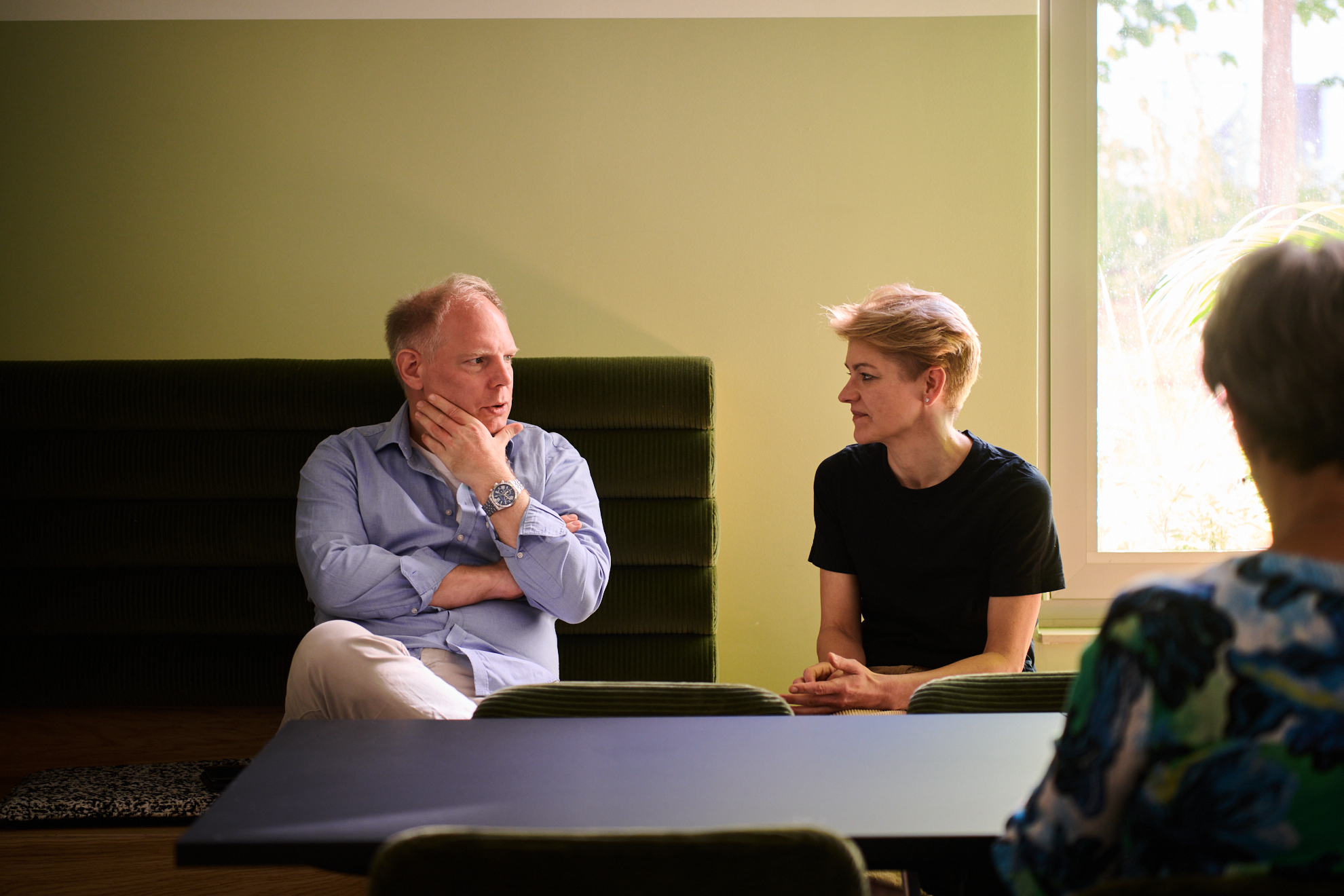 Zwei Personen im Gespräch in einem modernen Raum mit grünen Wänden und einem Tisch.