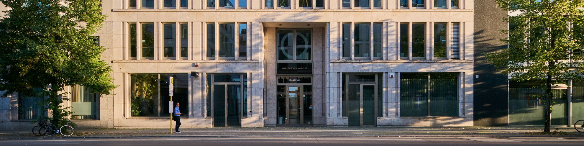 Moderne Gebäudeansicht mit großen Fenstern, Eingangsbereich und Bäumen im Vordergrund.