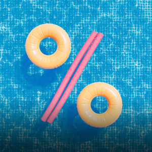 Zwei orangefarbene Schwimmreifen und ein pinker Schwimmstab bilden das Prozentzeichen auf blauem Wasserhintergrund.