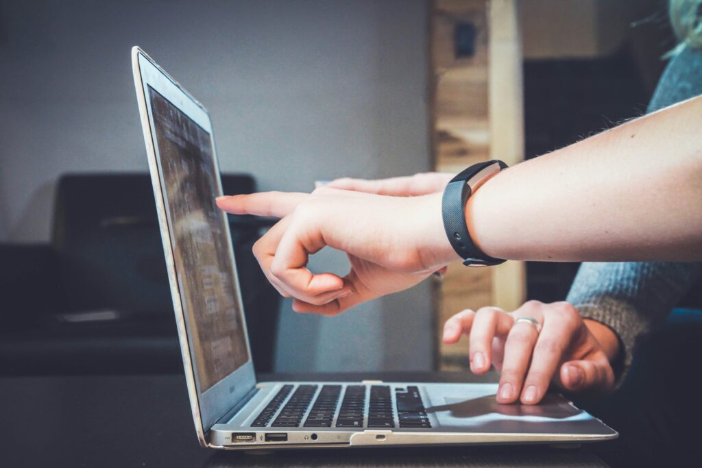 Eine Hand zeigt auf den Bildschirm eines Laptops, während eine andere Hand darauf tippt.